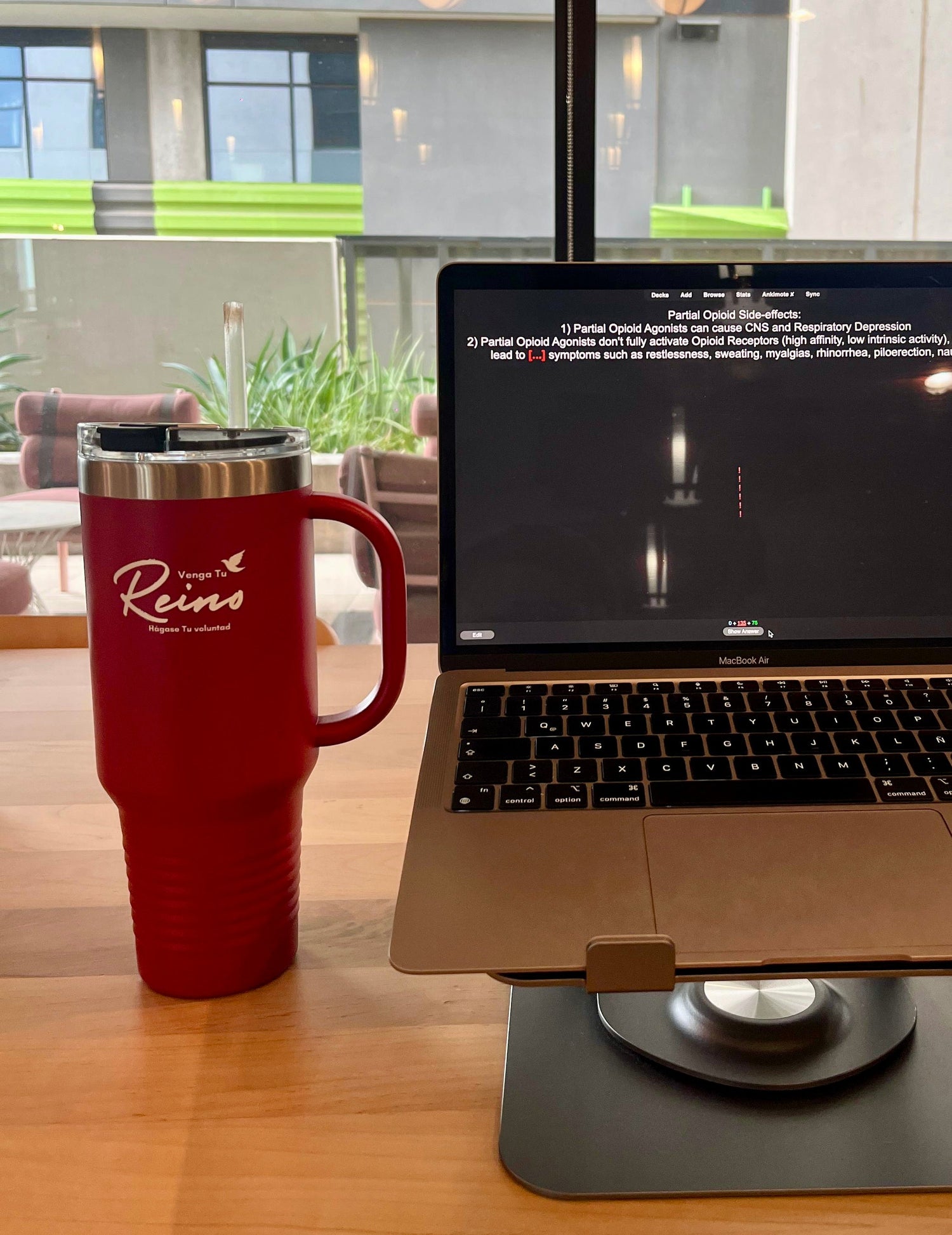 Red Christian travel mug with Spanish faith text on wooden desk next to laptop, modern indoor setting