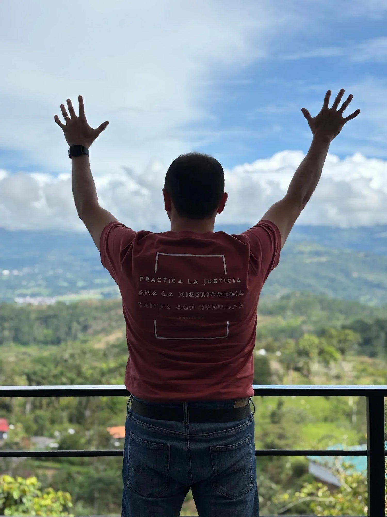 Man in Christian t-shirt with arms raised, overlooking scenic valley and cloudy sky
