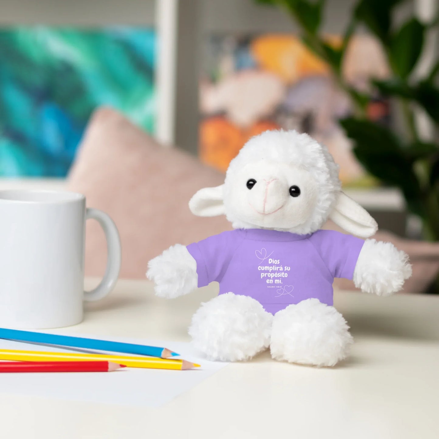 Plush lamb in purple Christian shirt with Spanish faith quote on desk, next to mug and colored pencils.