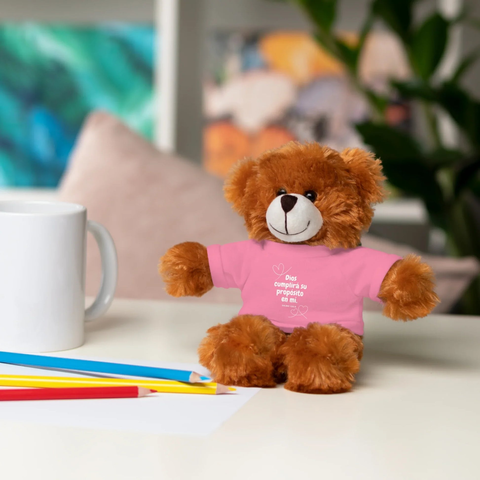 Plush teddy bear in pink shirt with Spanish faith message, on desk with pencils and mug