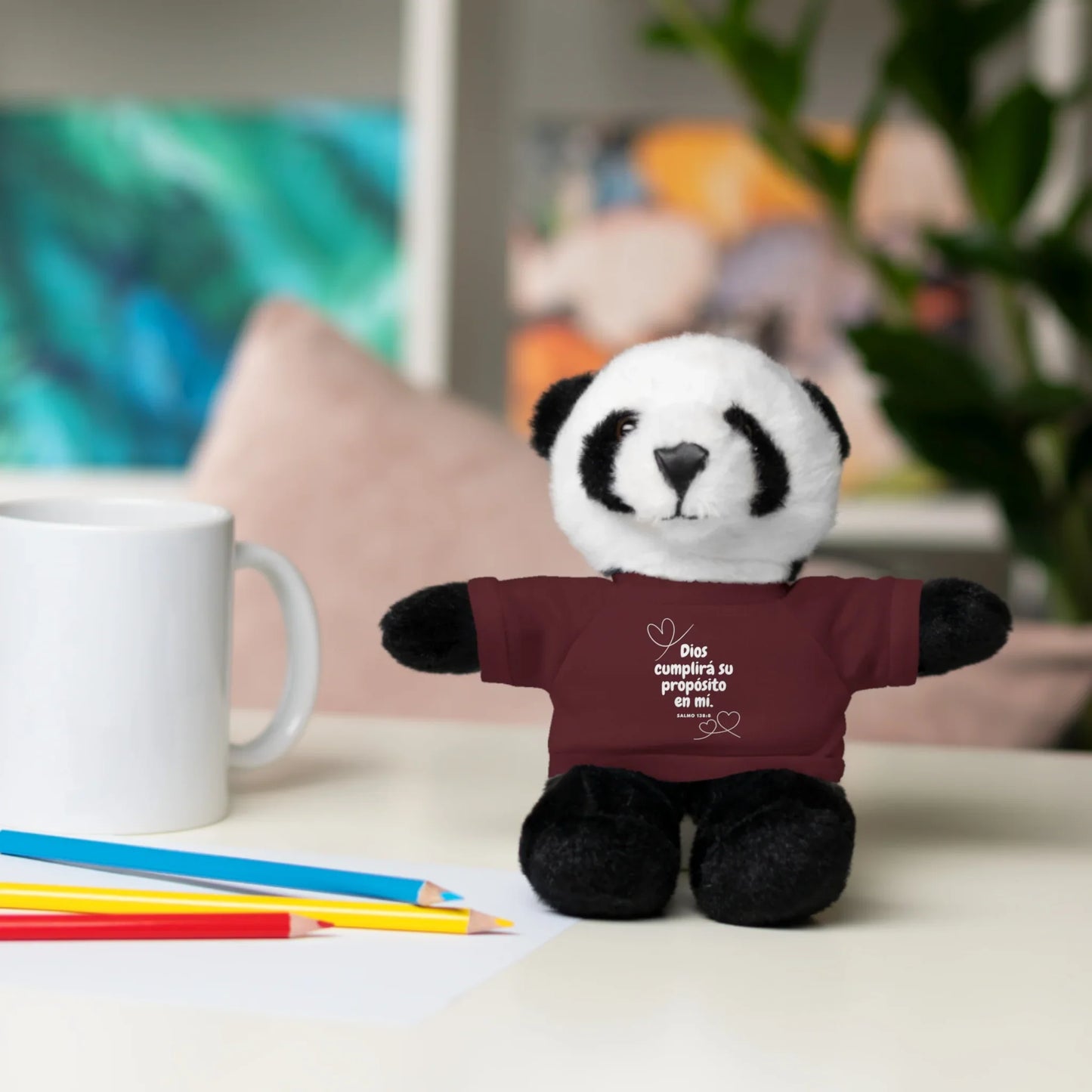 Plush panda toy in maroon shirt with Spanish Christian message, on desk with mug and pencils