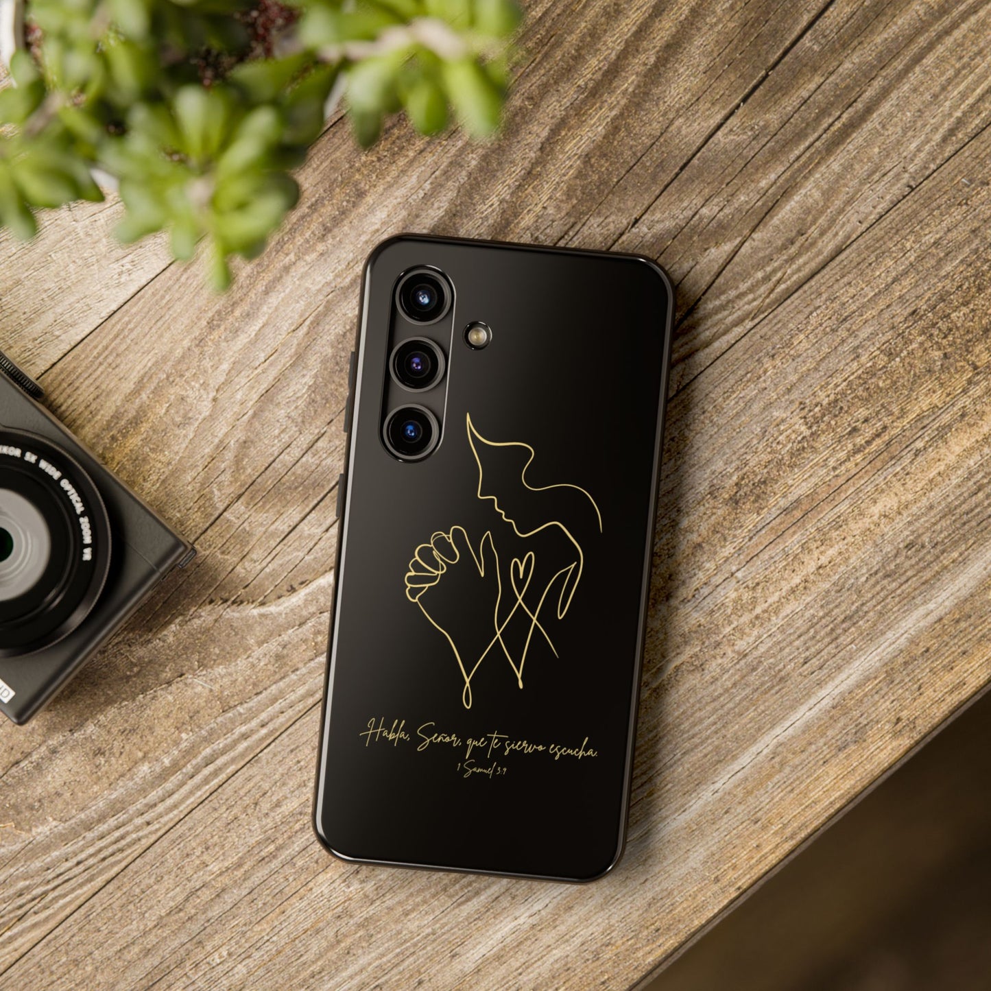 Black phone case with minimalist gold praying figure and Spanish Bible verse, on wooden table