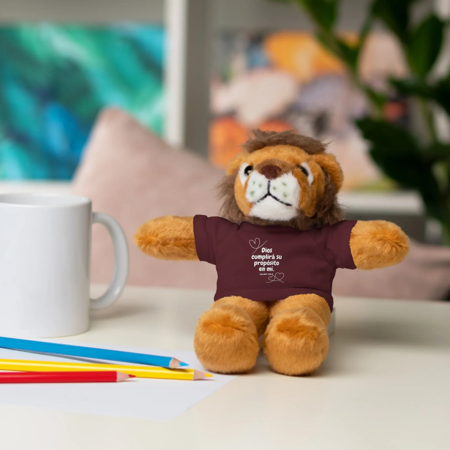 Plush lion toy with Christian quote shirt on desk, next to mug and colored pencils