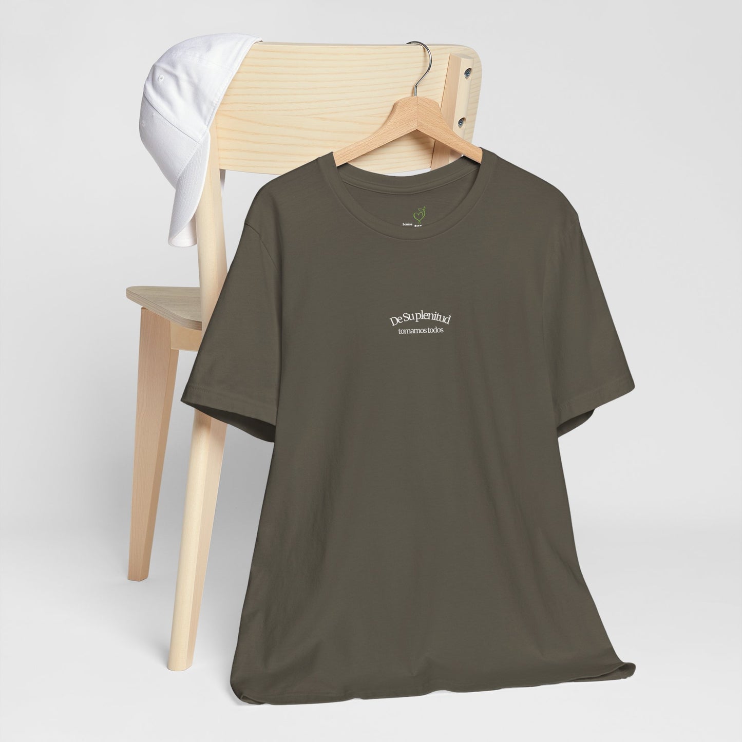 Olive green Christian t-shirt with Spanish faith message on hanger, white cap on wooden chair