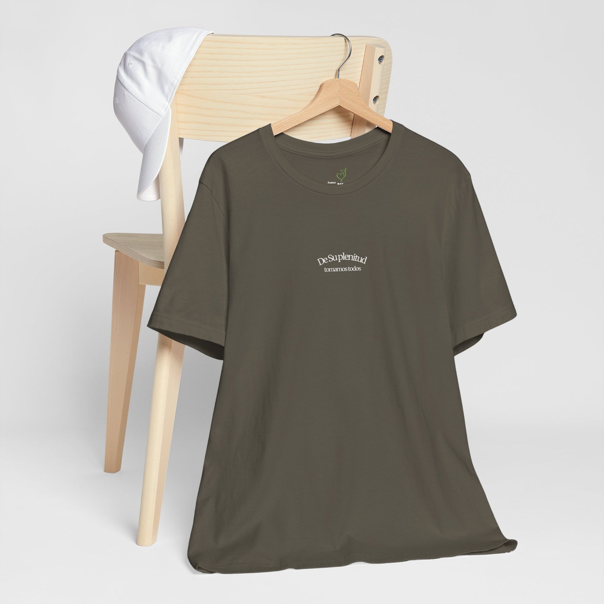 Olive green Christian t-shirt with Spanish faith message on hanger, white cap on wooden chair