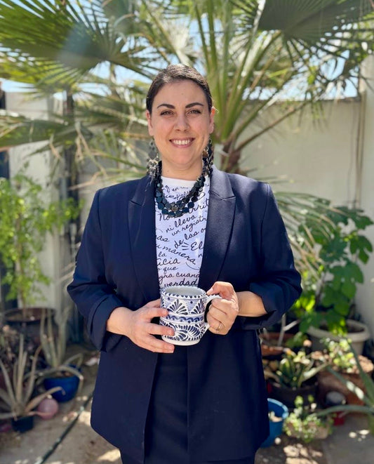 Smiling woman in navy blazer holding patterned coffee mug, standing in garden with palm trees
