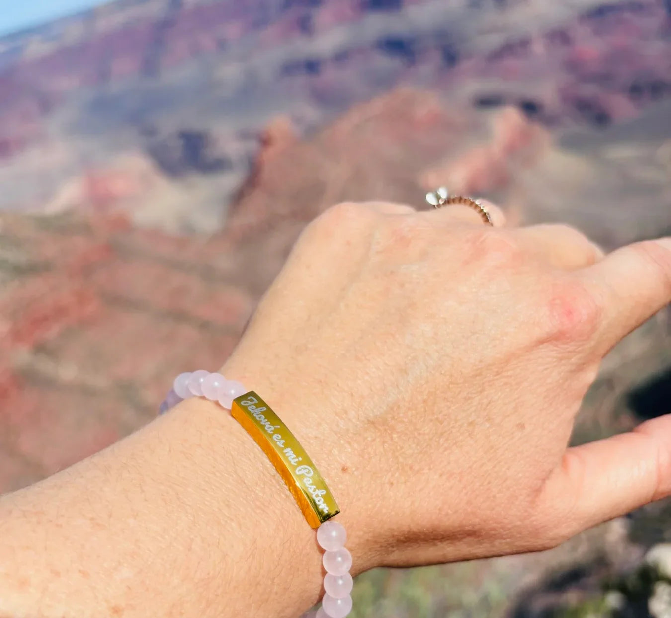 Pulsera cristiana rosa con placa dorada grabada 'Jehová es mi Pastor' en paisaje natural