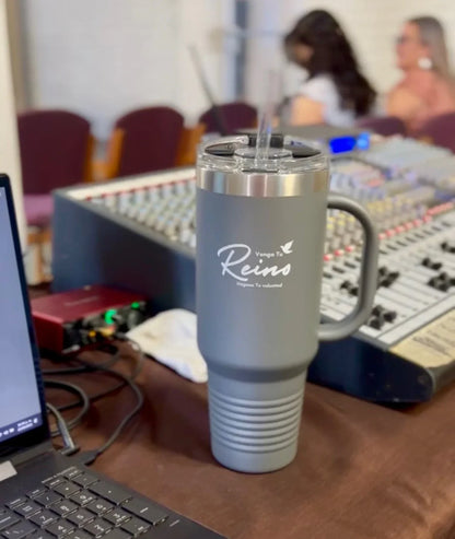Gray Christian tumbler with "Venga Tu Reino" design on a table near audio mixers and a laptop