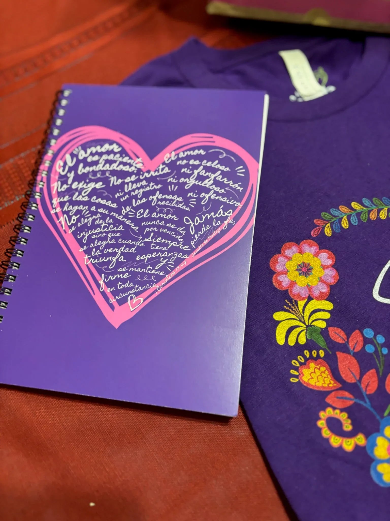 Purple Christian notebook with heart and Spanish scripture, next to floral shirt on red fabric.