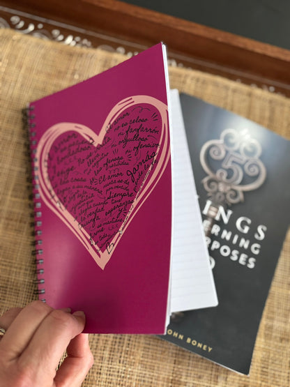 Hand holding a pink heart-themed Christian notebook with Spanish script, book in background