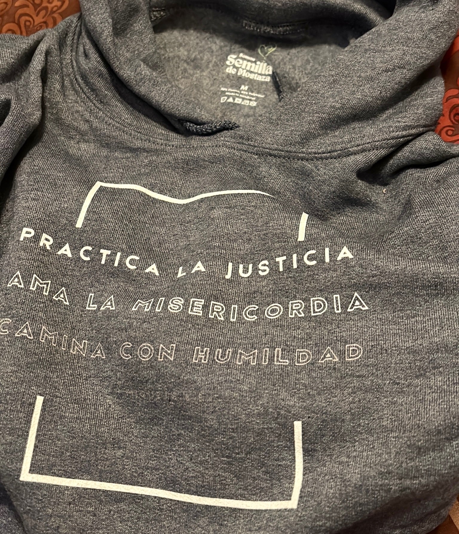 Gray Christian hoodie with Spanish inspirational text 'Practica la justicia, ama la misericordia, camina con humildad' on front.