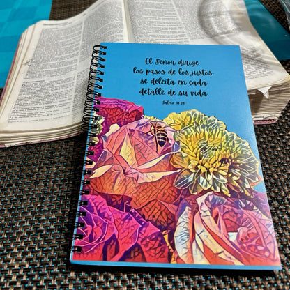 Colorful Christian floral notebook with Spanish Bible verse beside open Bible on table