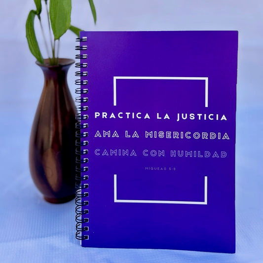 Purple Christian spiral notebook with Spanish Bible verse, beside a vase with green leaves.