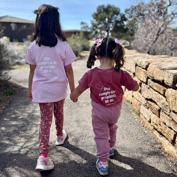 Two young girls wearing Christian faith t-shirts with Spanish scripture, holding hands outdoors