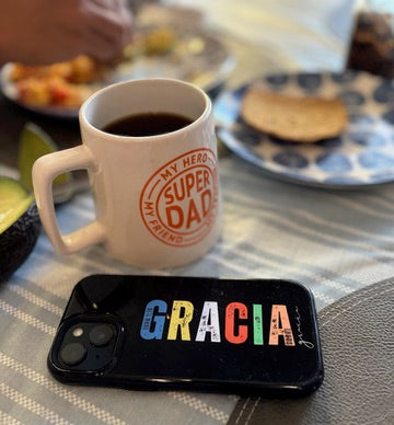 Coffee cup with 'Super Dad' text and phone with colorful 'GRACIA' case on breakfast table