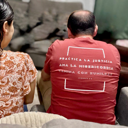 Man wearing red Christian t-shirt with Spanish Bible verse, sitting indoors on sofa
