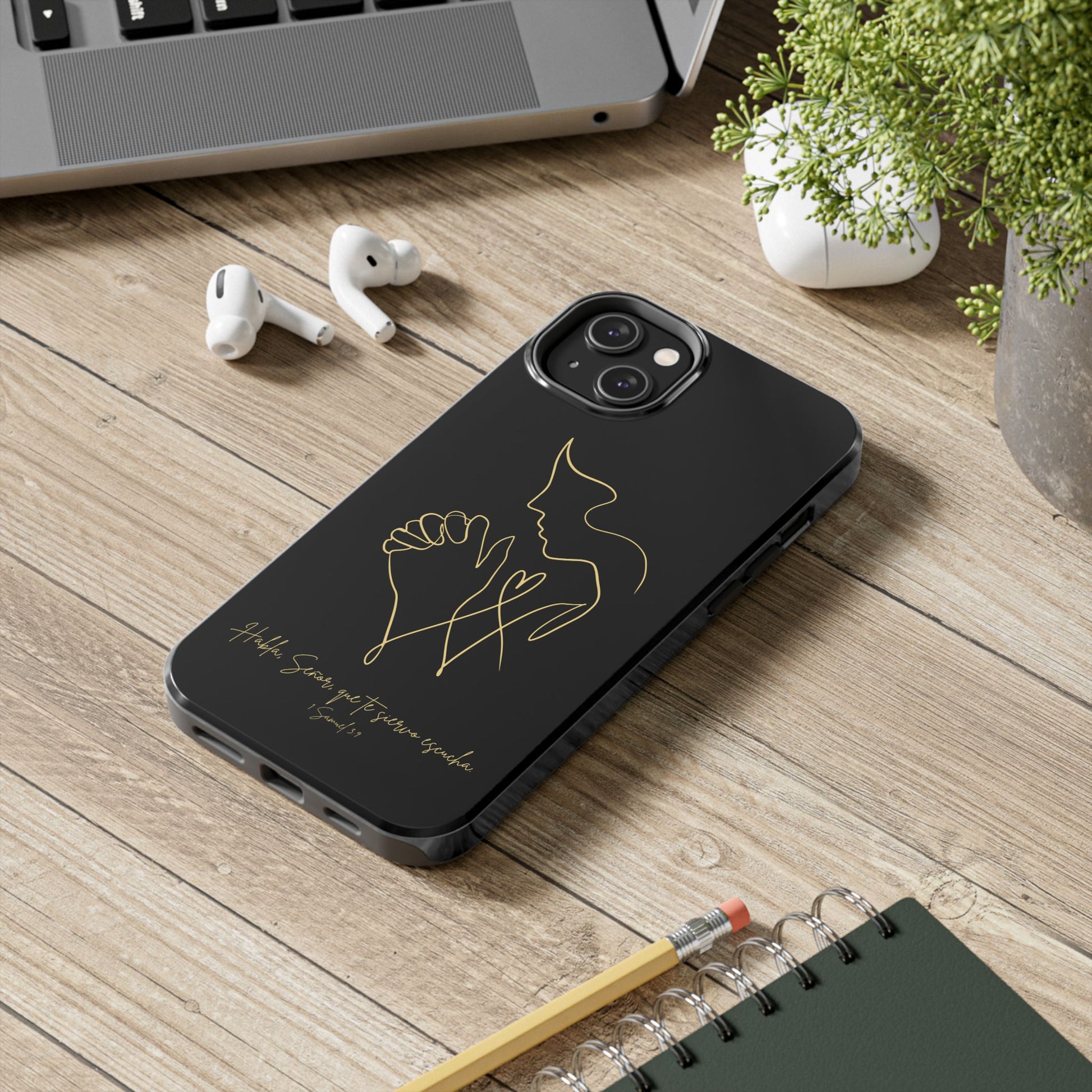 Black phone case with gold line art of praying hands and a heart, placed on a desk.