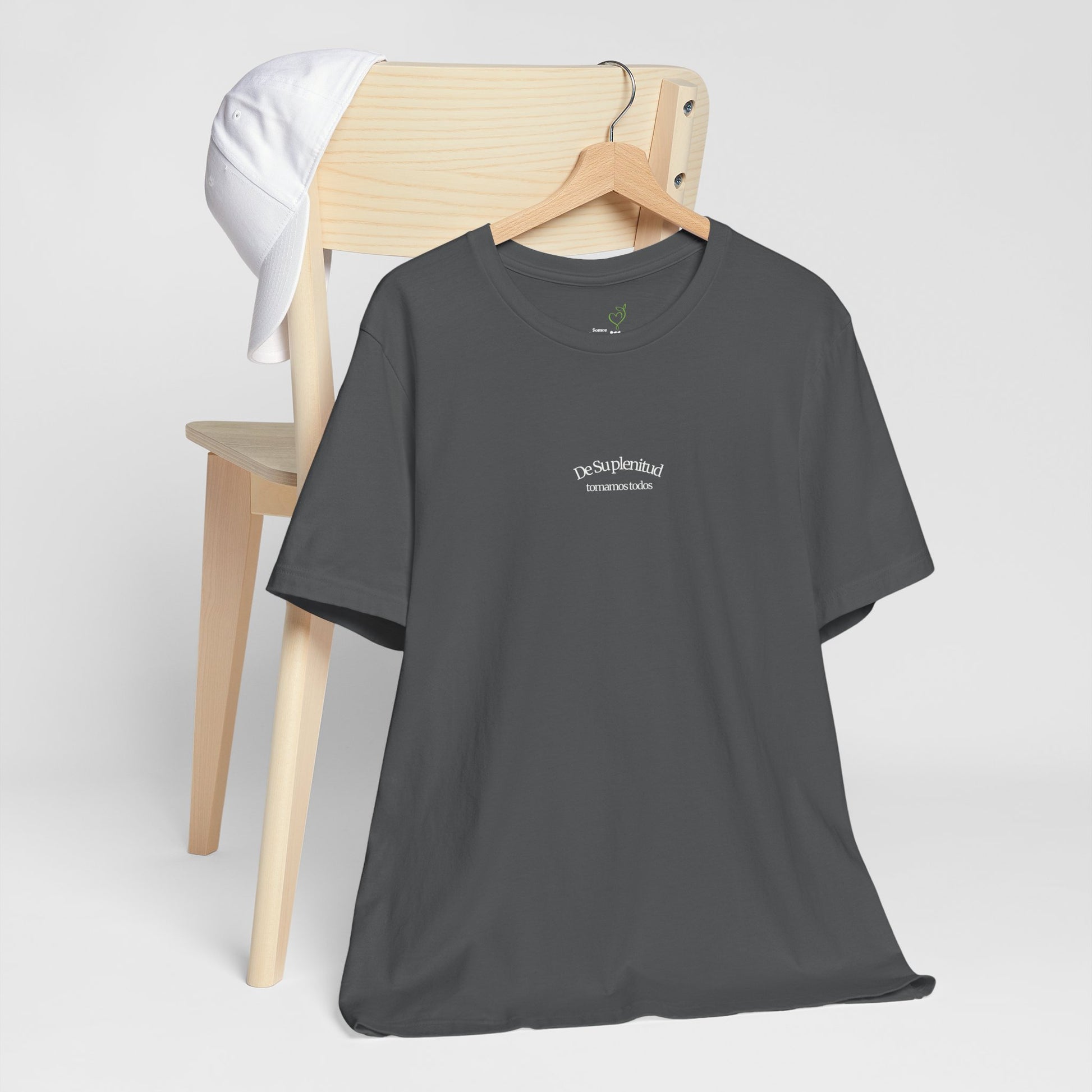 Charcoal Christian t-shirt with Spanish faith message on hanger, white cap on wooden chair