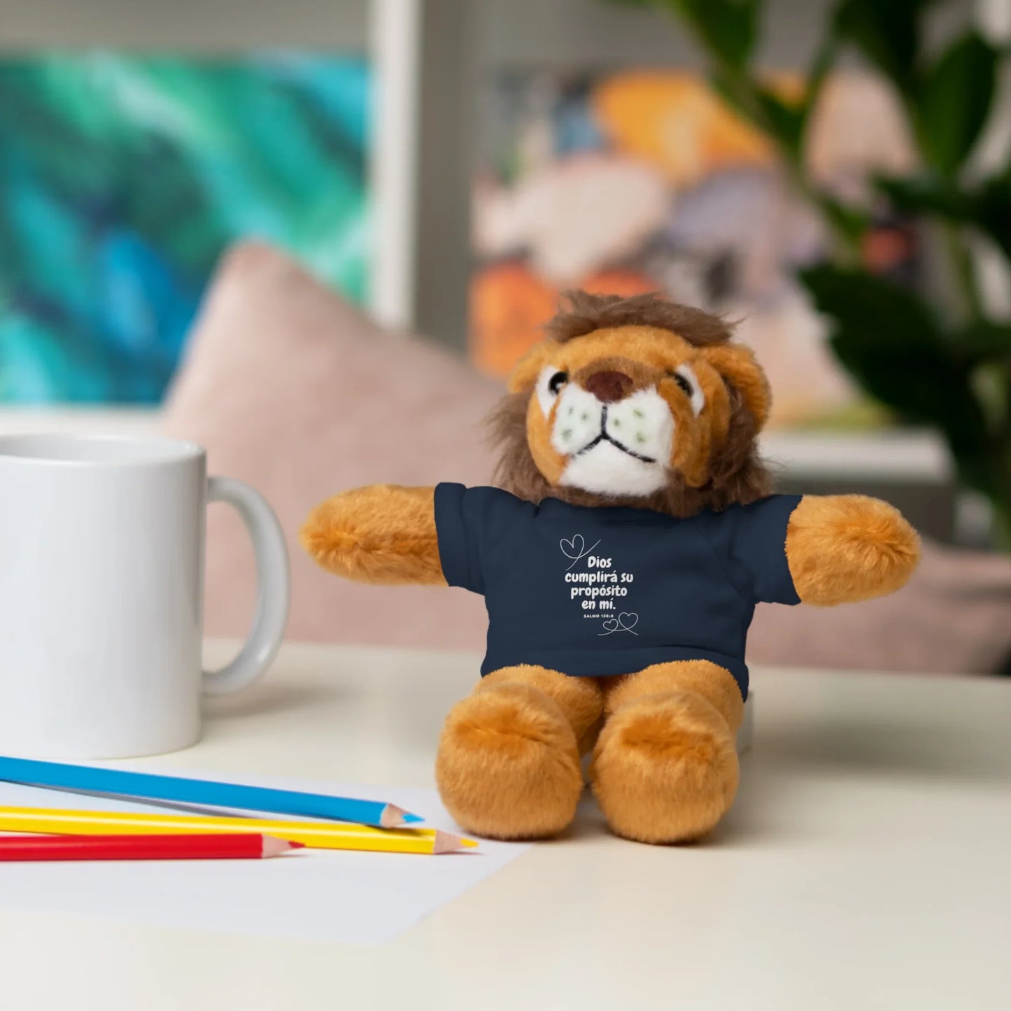 Plush lion toy wears Christian message shirt, on desk with mug and colored pencils.