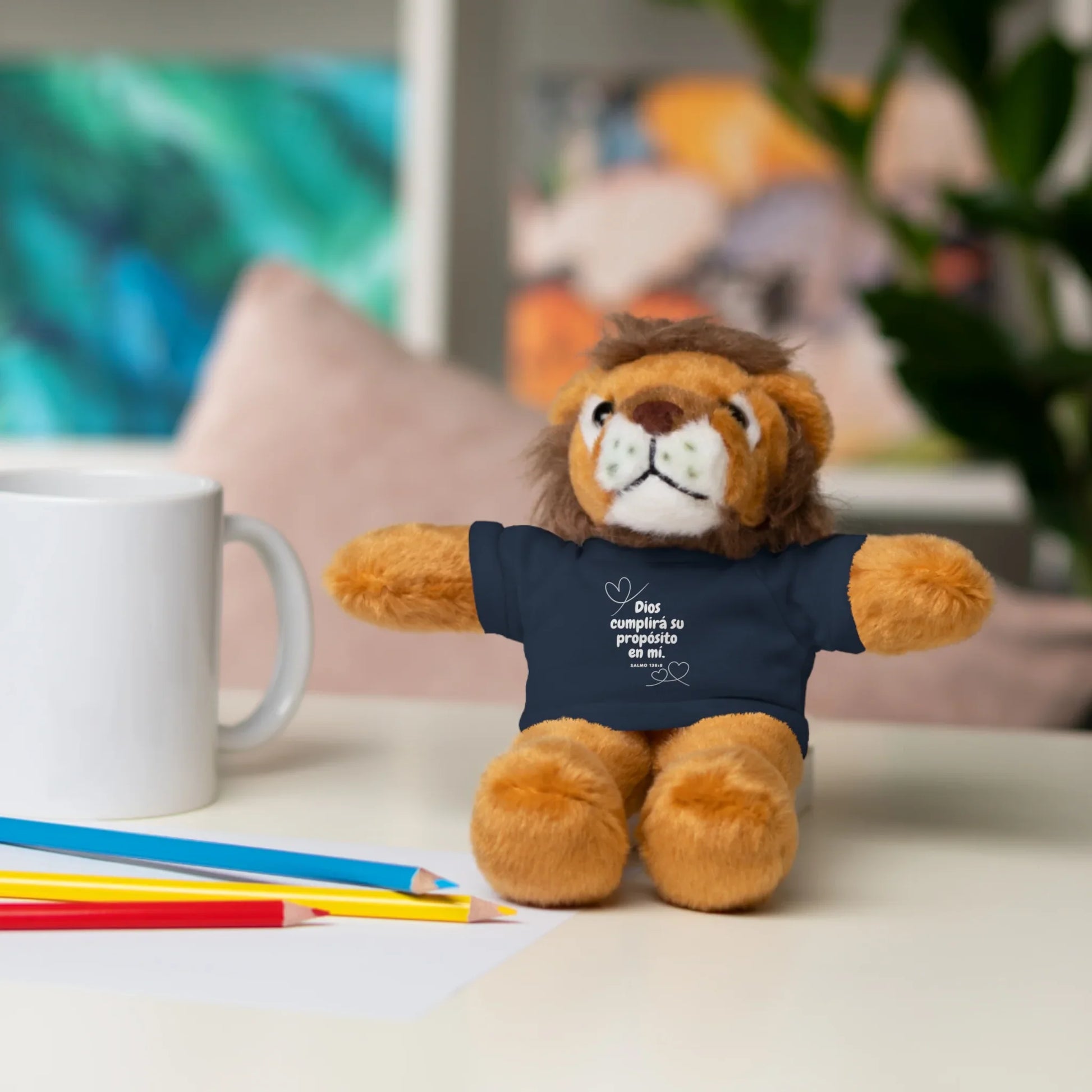 Plush lion toy wears Christian message shirt, on desk with mug and colored pencils.