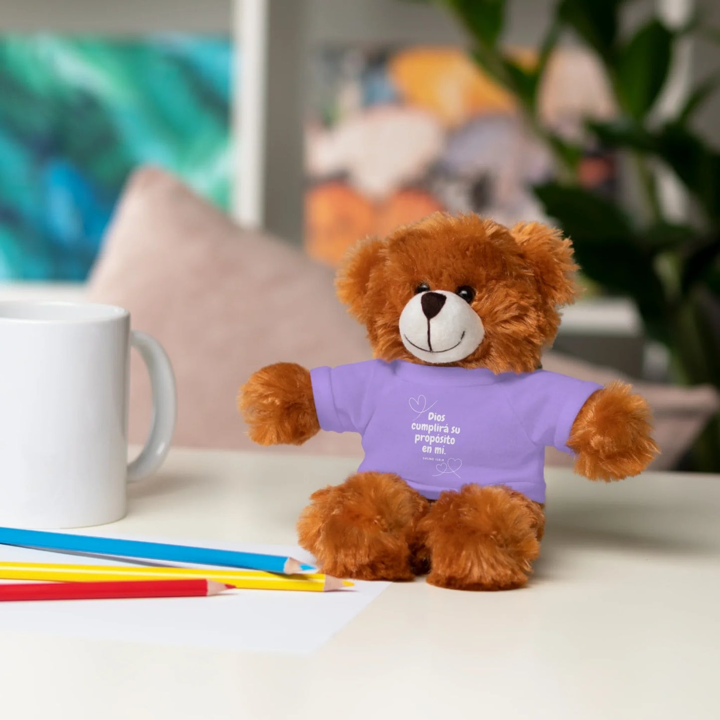 Plush teddy bear in a purple Christian T-shirt with Spanish text on a desk with pencils and mug.