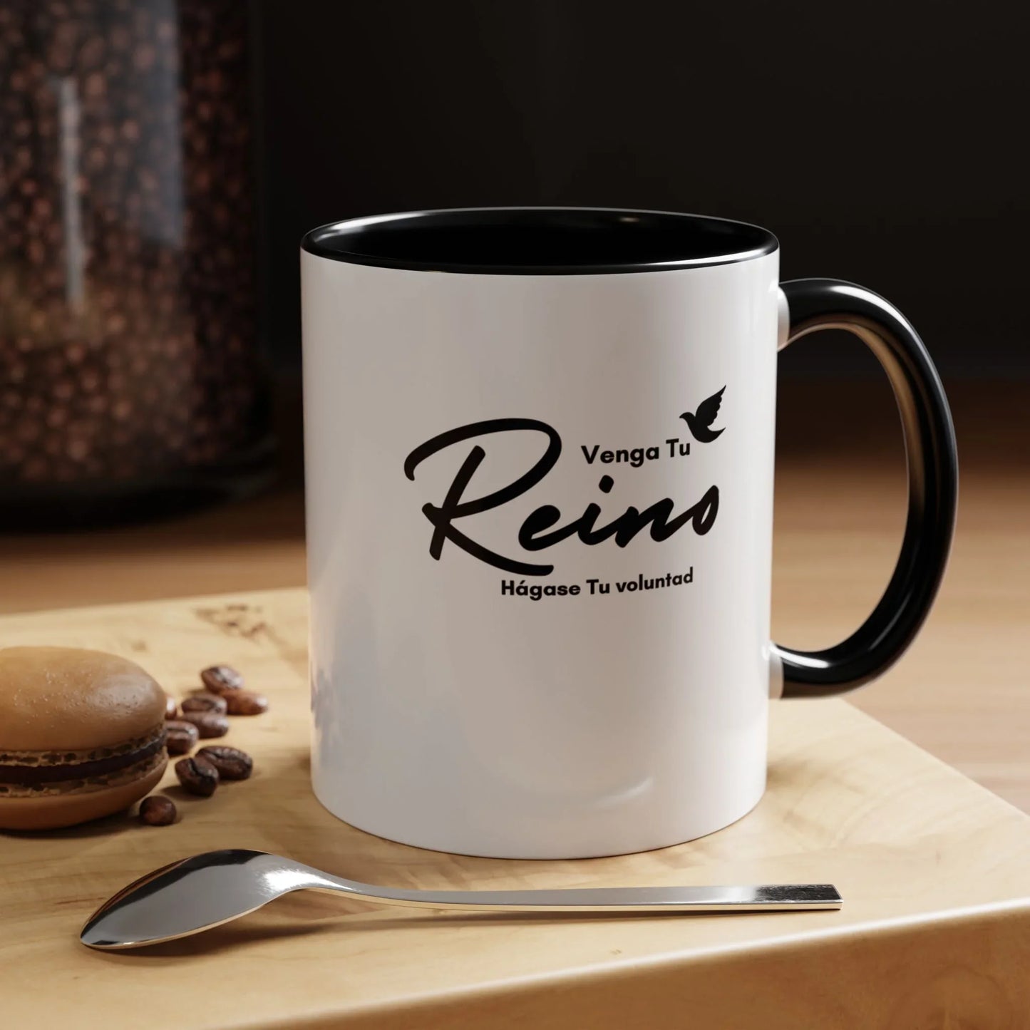 White Christian mug with Spanish Scripture text, black handle, spoon, coffee beans and macaron nearby