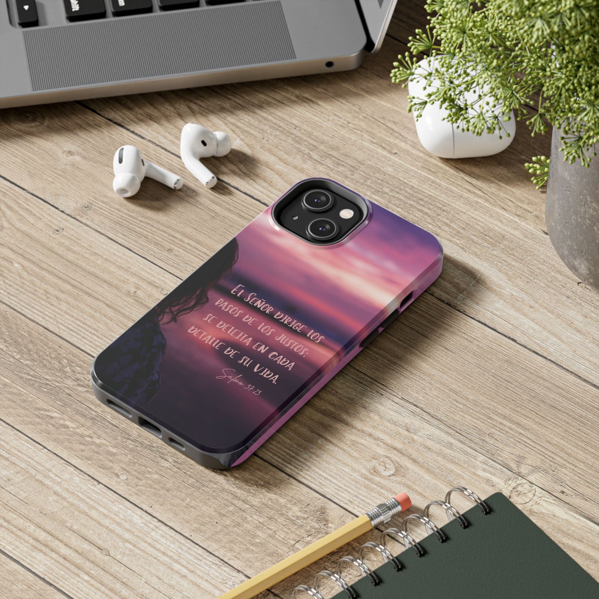 Christian phone case with Spanish Scripture on desk with laptop, plant, AirPods, and notebook.