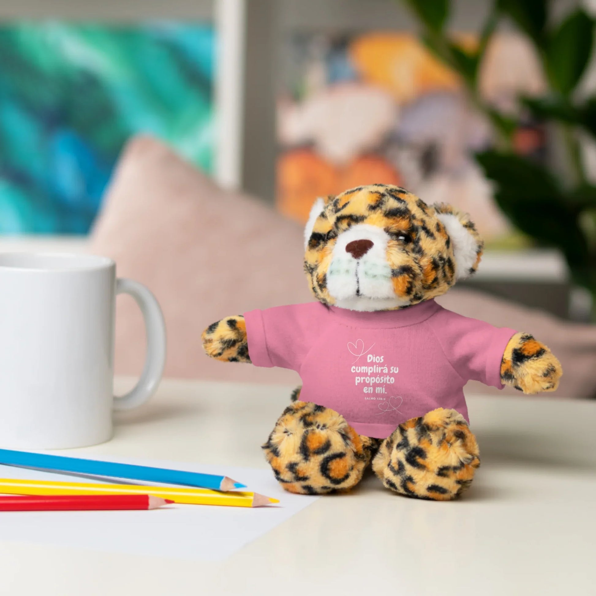 Leopard plush toy with pink Christian message shirt, mug, and colored pencils on desk