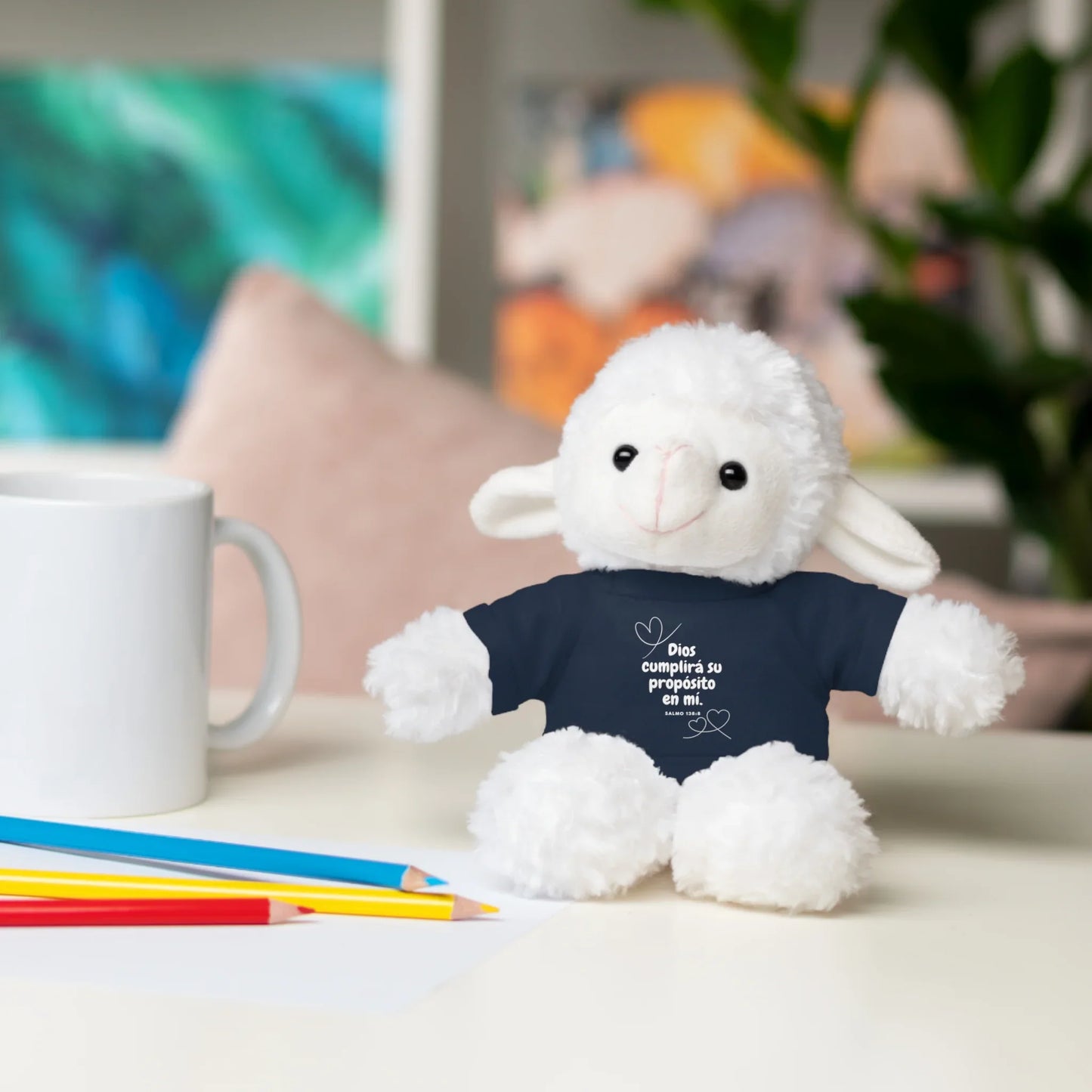 Plush lamb toy wearing a Christian message shirt on a desk with mug and colored pencils
