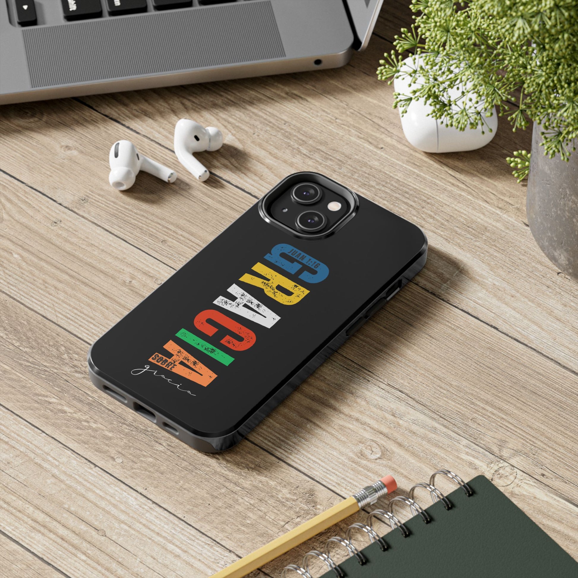 Black phone case with colorful Christian GRACIA text design on wooden desk beside laptop, AirPods, and plant