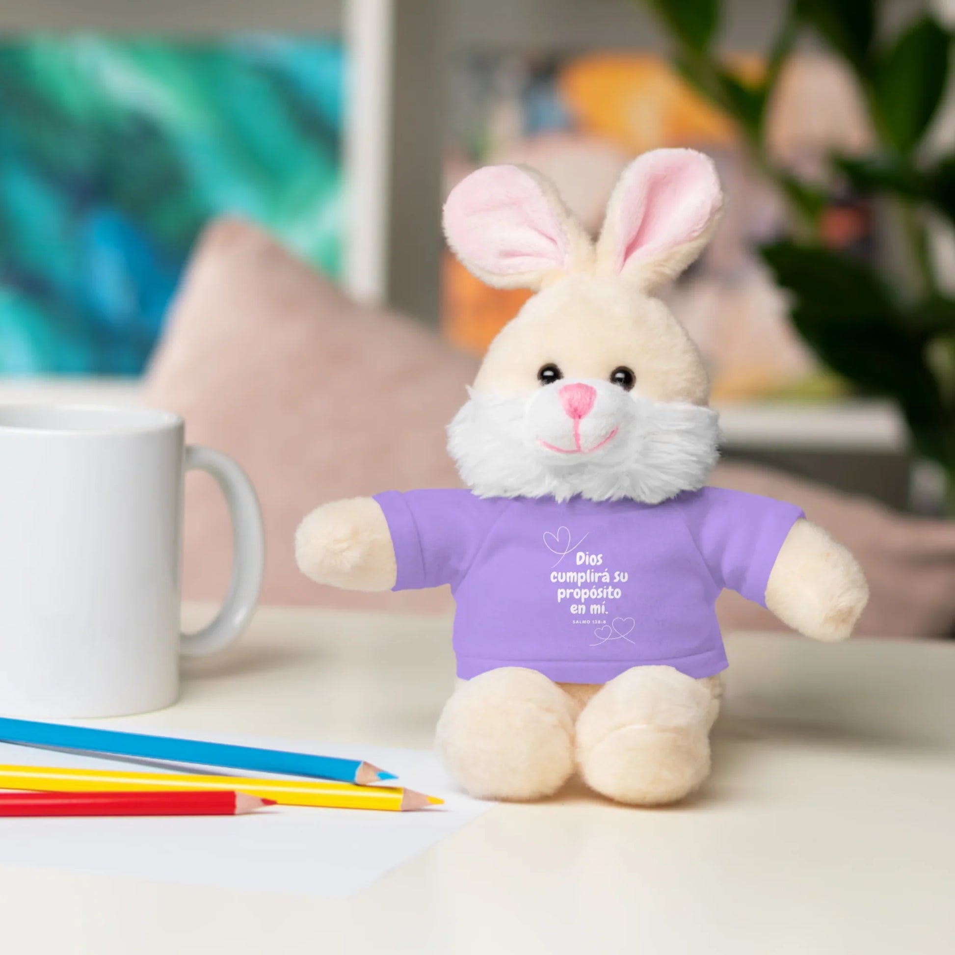 Plush bunny in purple shirt with Christian message, on a desk with mug and colored pencils