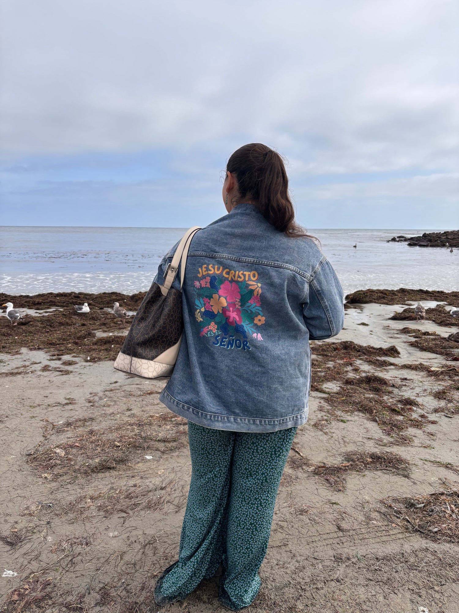 woman on the beach in a denim jacket with a colorful "Jesucristo es el Señor" design